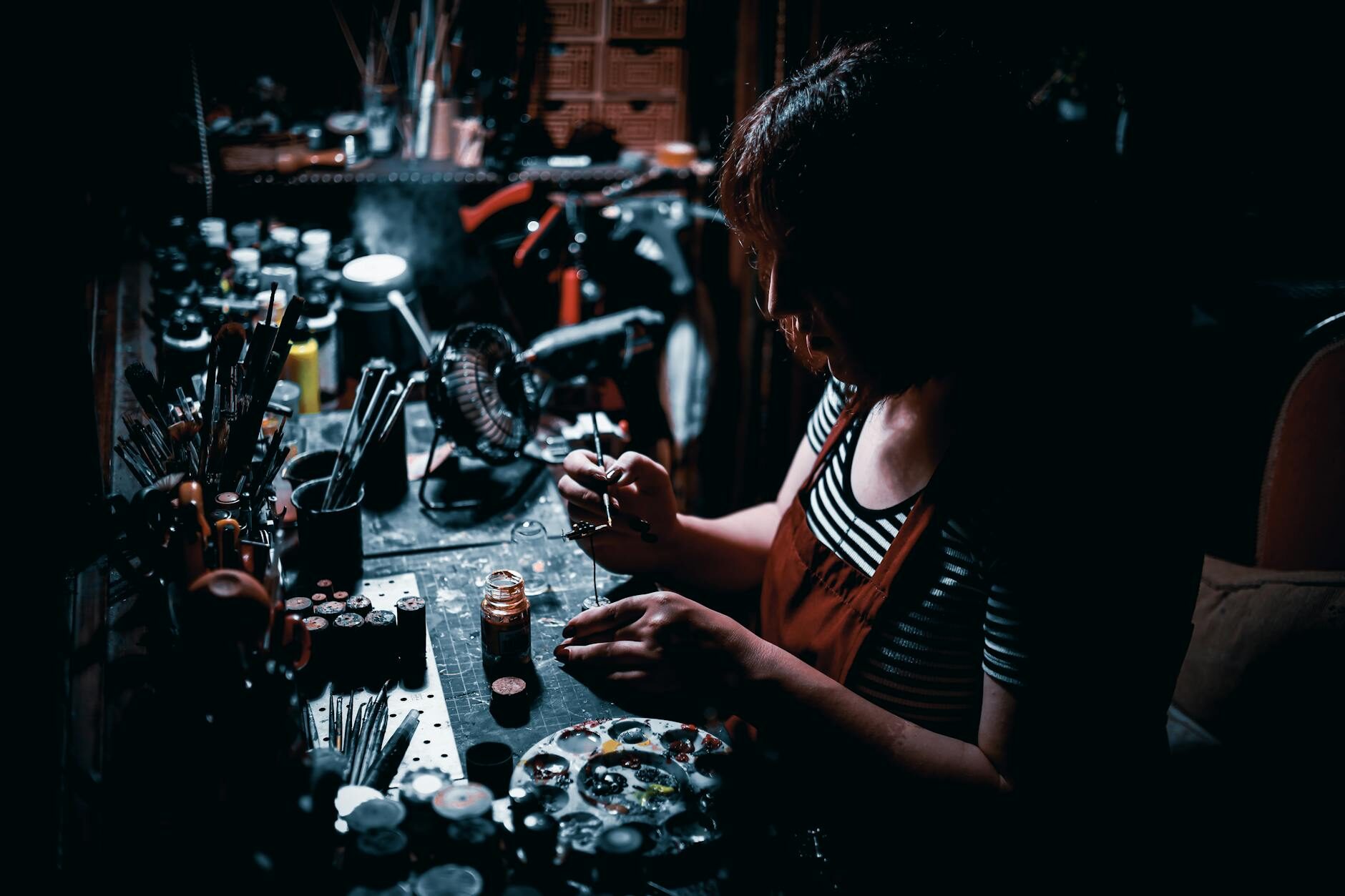 Artist working at a bench in a dramatically lit dark workshop surrounded by brushes and paints